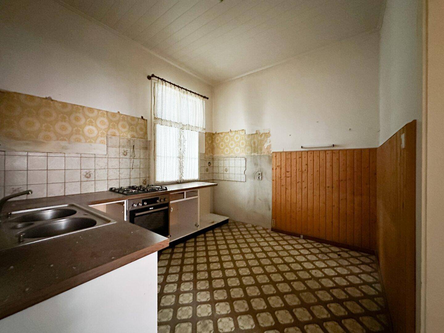 Küche in renovierungsbedürftigem Zustand mit Gasherd, Spüle und gemusterten Wand- und Bodenfliesen.