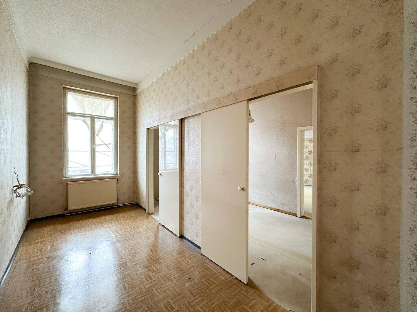 Geräumiger Raum mit Fenster, Heizkörper und alten Tapeten, bereit für eine umfassende Renovierung.