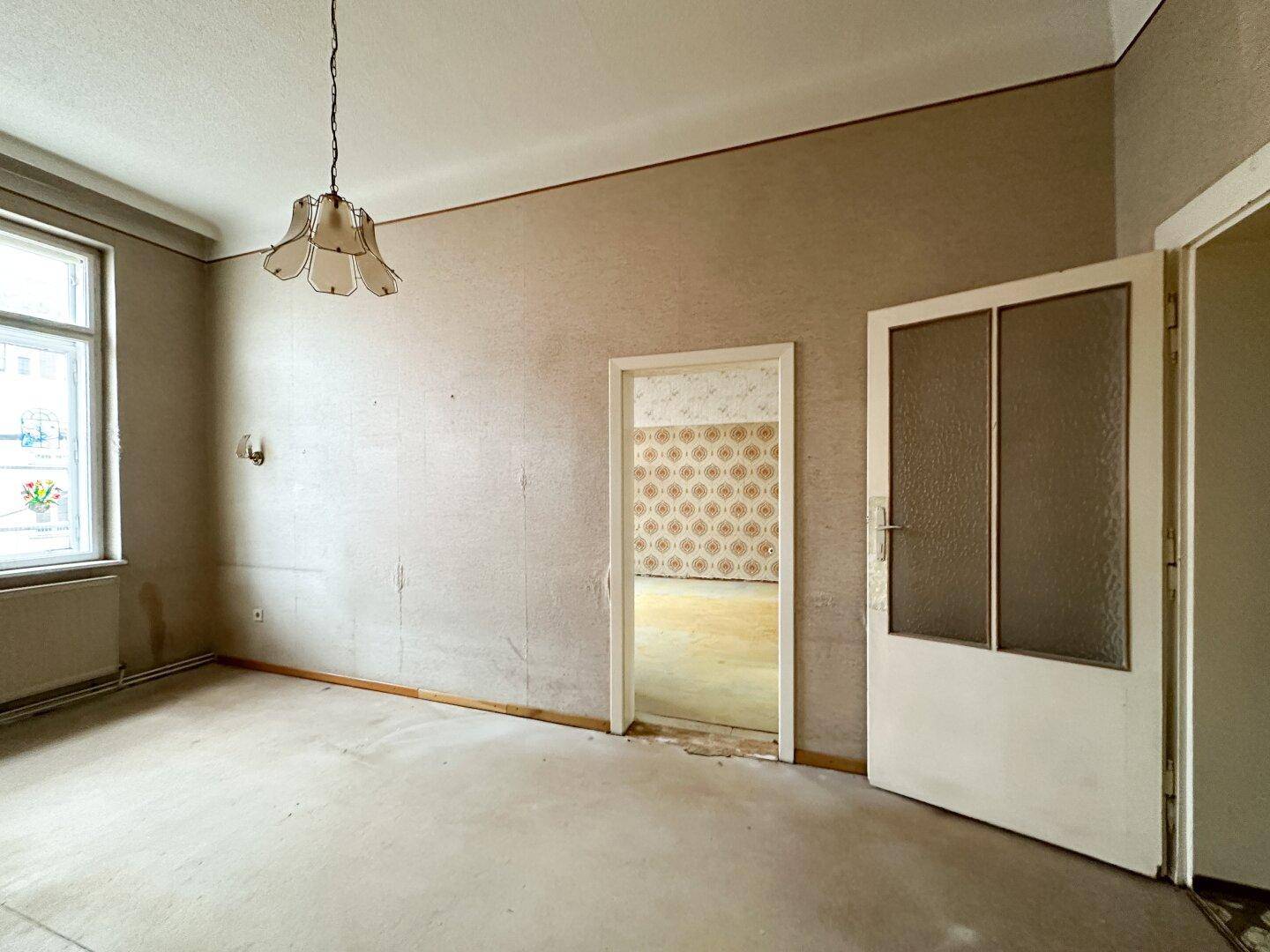 Leerer Raum mit Fenster, Heizkörper und einer alten Deckenleuchte, bereit zur Neugestaltung und Modernisierung.