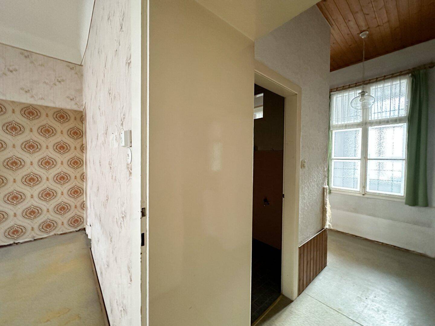Flur mit Holzdecke und Blick in einen angrenzenden Raum mit gemusterter Tapete, der renoviert werden muss.
