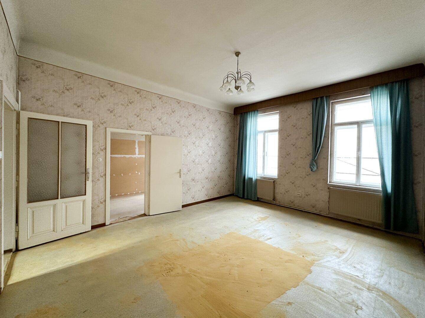 Geräumiger Wohnbereich mit zwei Fenstern, Heizkörpern und veralteten Tapeten, die erneuert werden müssen.