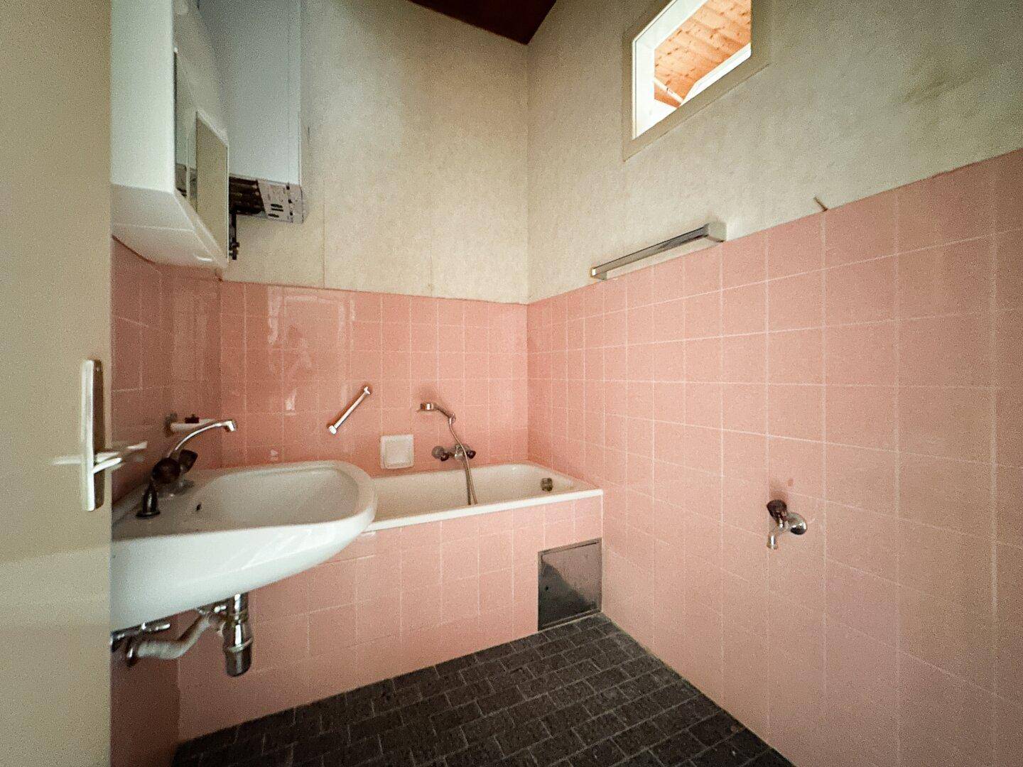 Badezimmer mit rosa Fliesen, Badewanne, Waschbecken und einem kleinen Oberlichtfenster, im Stil der 80er Jahre.