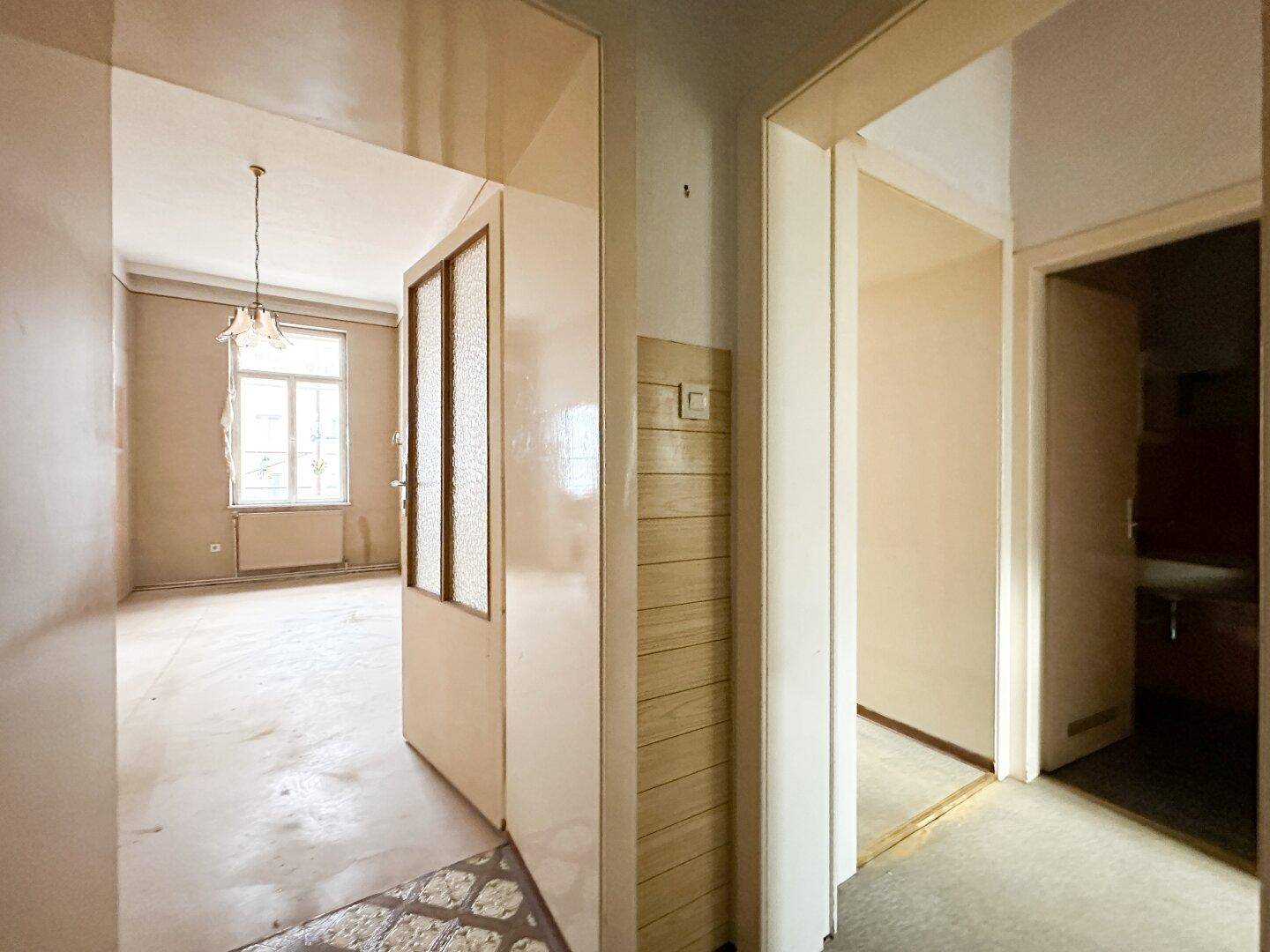 Flur mit Blick in einen leeren Raum, der auf Renovierung wartet, mit alter Tür und Holzverkleidung.