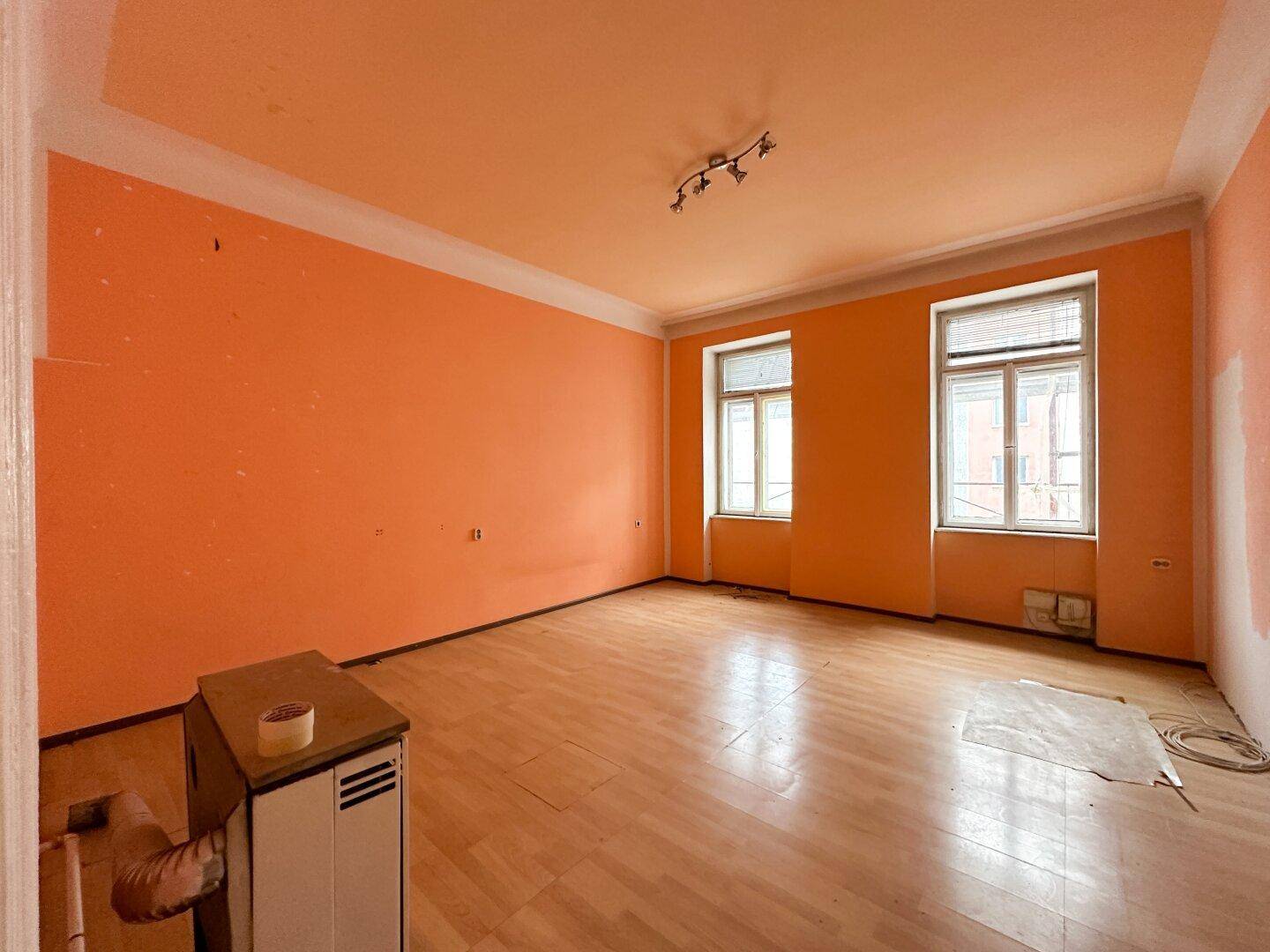 Geräumiges, leeres Zimmer mit leuchtend orangefarbenen Wänden, Parkettboden und zwei Fenstern.