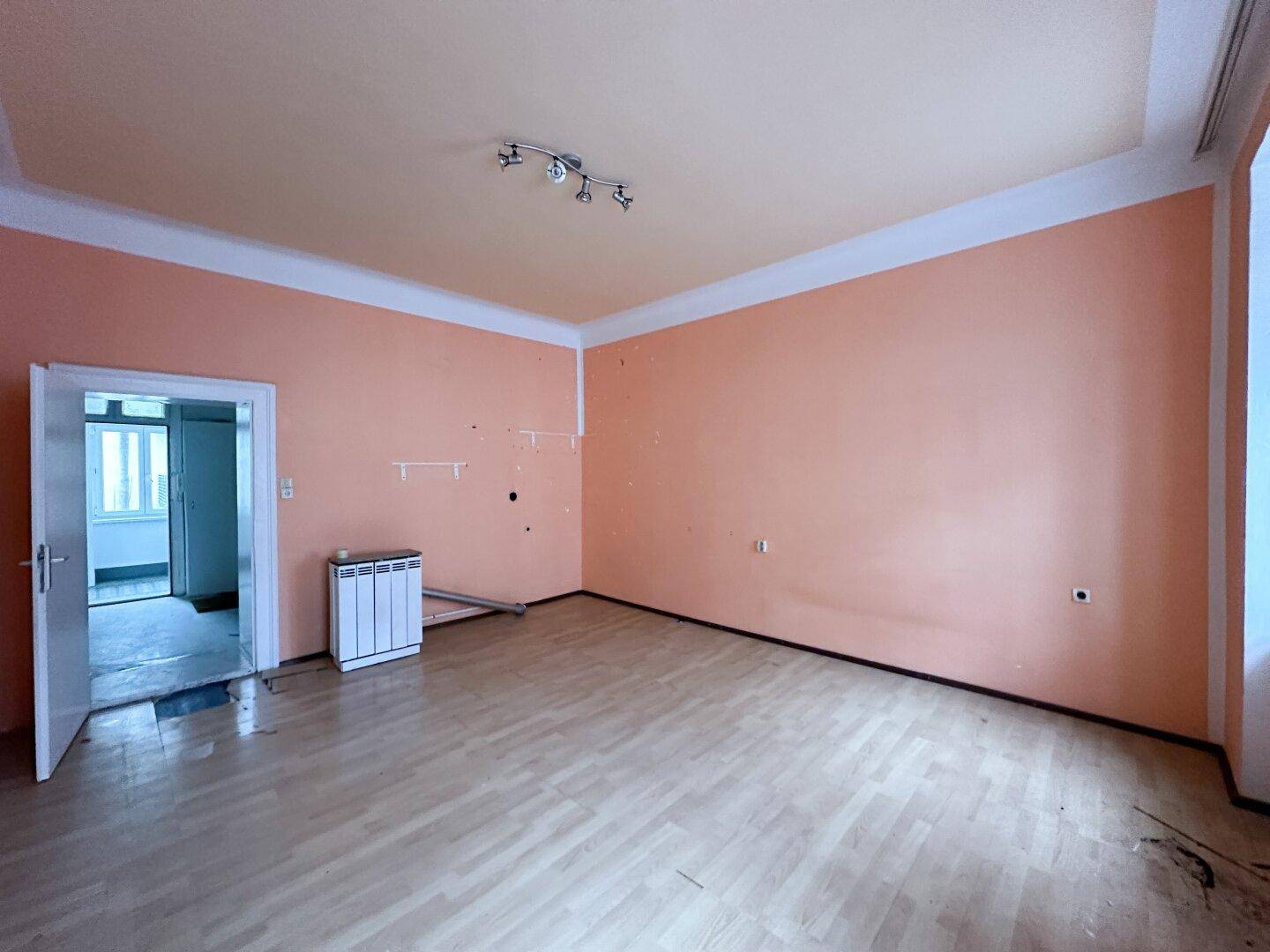 Helles, leeres Zimmer mit orangefarbenen Wänden, Parkettboden und offener Tür zu einem weiteren Raum.