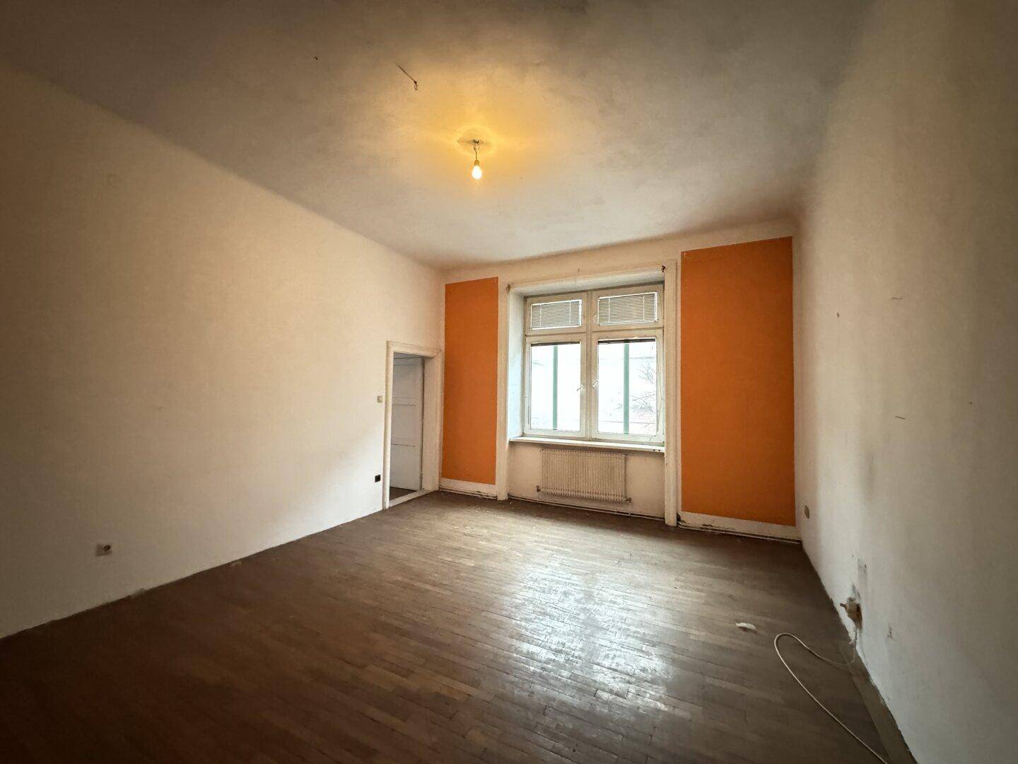 Geräumiges leeres Zimmer mit orangefarbenen Akzentwänden und abgenutztem Holzparkettboden.