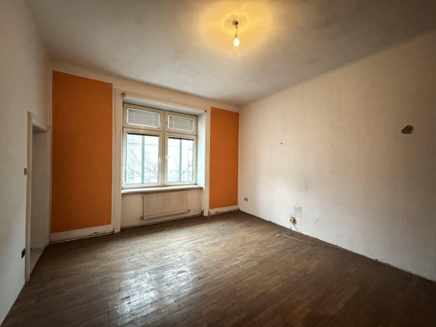 Helles Zimmer mit orangefarbenen Wänden, einem Fenster und einem renovierungsbedürftigen Parkettboden.