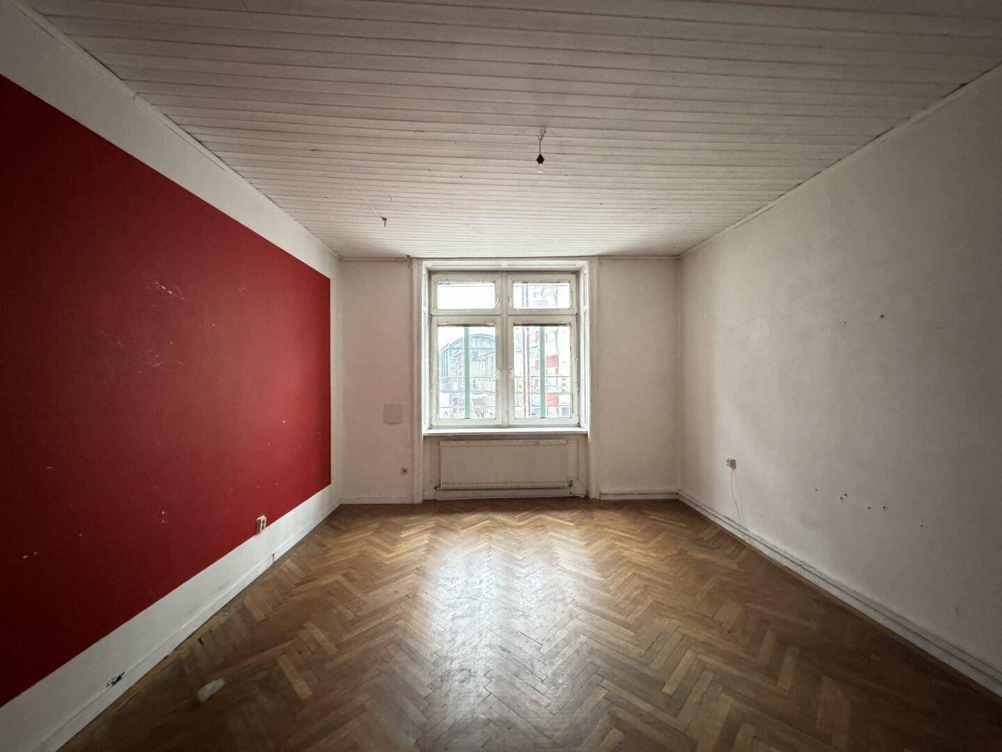Leeres Zimmer mit roter Akzentwand, Fischgrätparkett und einer Holzdeckenverkleidung.