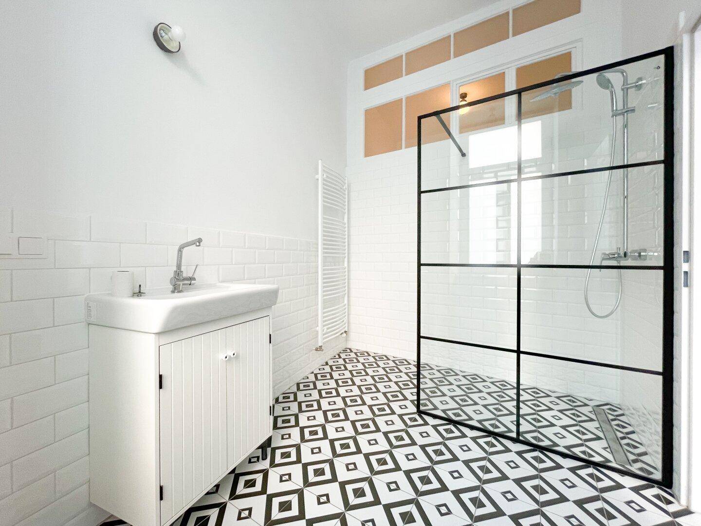 Helles Badezimmer mit moderner Duschkabine, geometrischem Fliesenboden und Fenster.
