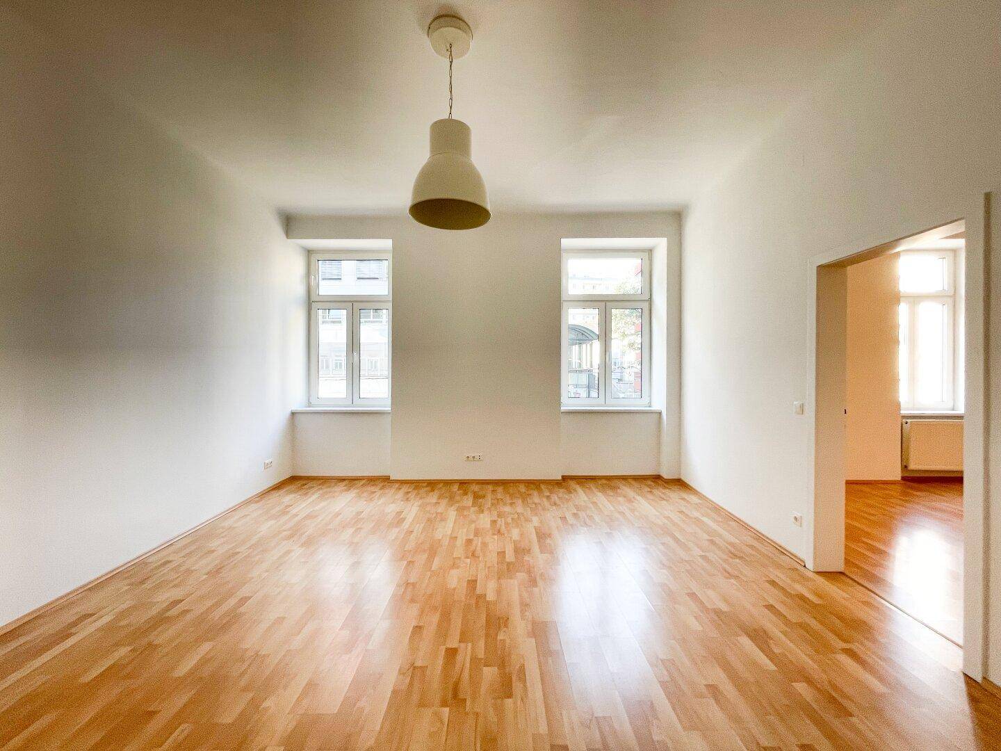 Geräumiges Wohnzimmer mit hellem Parkettboden, weißen Wänden und zwei großen Fenstern.