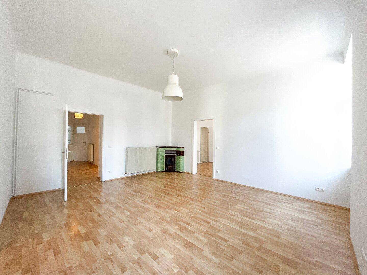 Helles Wohnzimmer mit Holzboden und einem traditionellen Kachelofen in der Ecke.
