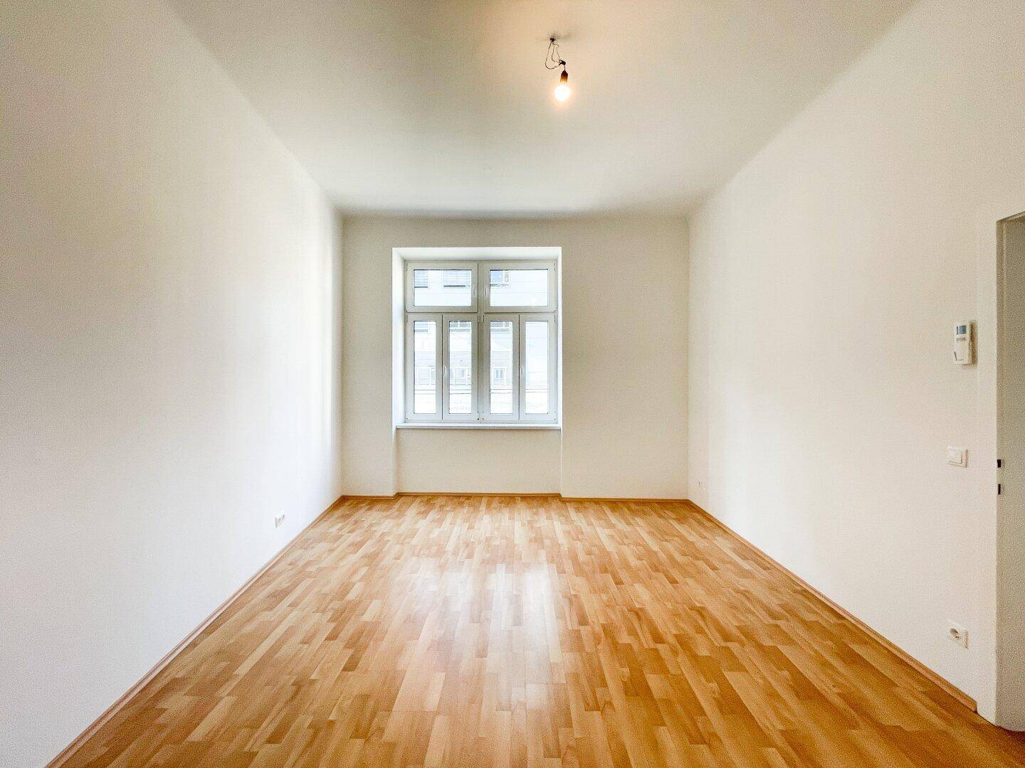 Leeres Schlafzimmer mit hellem Parkettboden, weiß gestrichenen Wänden und einem großen Fenster.