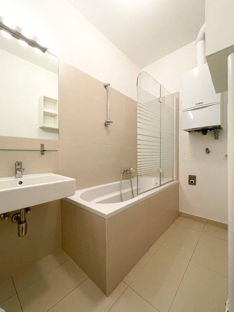 Funktionales Badezimmer mit Badewanne, Duschwand, Waschbecken und beigefarbenen Fliesen.