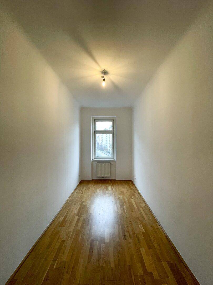 Schmales, helles Zimmer mit Parkettboden und einem Fenster am Ende des Raumes.