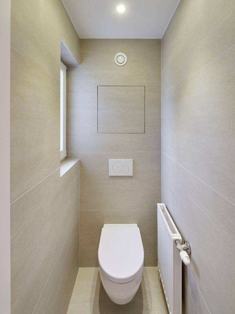 Modernes Gäste-WC mit Wand-WC, beigen Fliesen und kleinem Fenster.