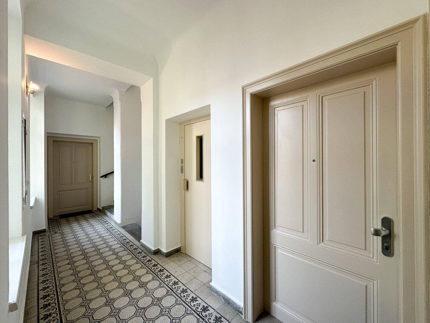 Heller Flur mit gemusterten Fliesenboden, hohen Decken und Zugang zum Aufzug in einem Altbau.