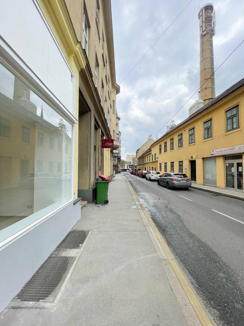 Blick auf eine städtische Straße mit parkenden Autos, historischen Gebäuden und einem markanten Industrieschornstein.