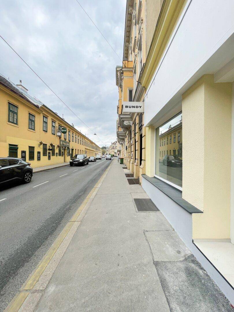 Eine ruhige Stadtstraße mit historischen Gebäuden, parkenden Autos und einem breiten Bürgersteig vor einem Geschäft.