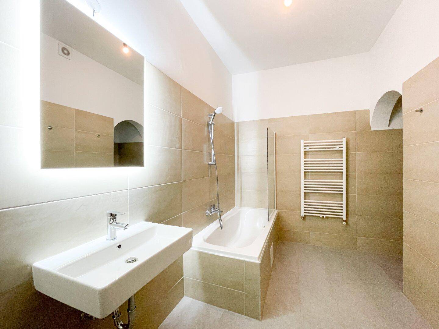 Modernes Badezimmer mit beigefarbenen Fliesen, Badewanne mit Duschfunktion und beleuchtetem Spiegel.