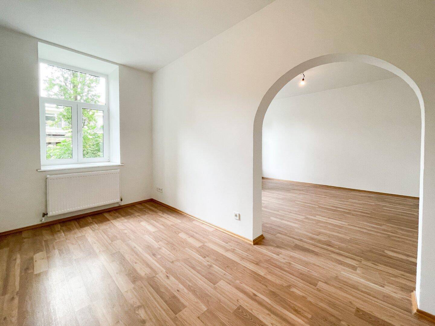 Heller Raum mit Holzboden und einem Fenster, das einen Blick ins Grüne bietet, verbunden durch einen Rundbogen.