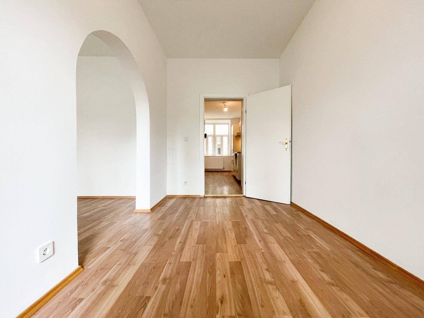 Leeres Zimmer mit Holzboden und offener Tür, die den Blick in eine angrenzende Küche freigibt.