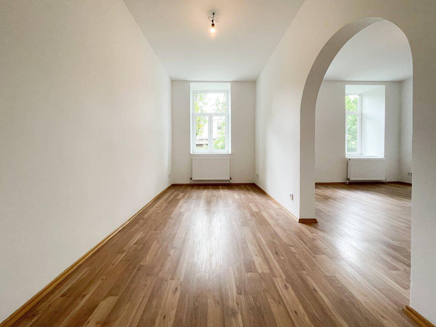 Langes, helles Zimmer mit Holzboden und einem Fenster, ein Rundbogen führt zu einem weiteren Bereich.
