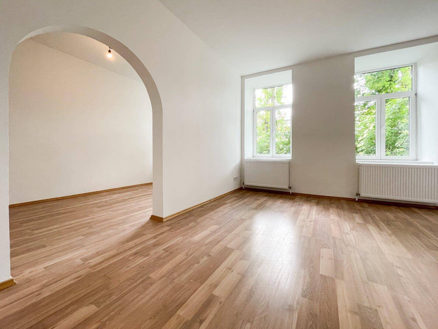 Helles, leeres Zimmer mit Holzboden und zwei Fenstern, verbunden durch einen Rundbogen zu einem weiteren Raum.