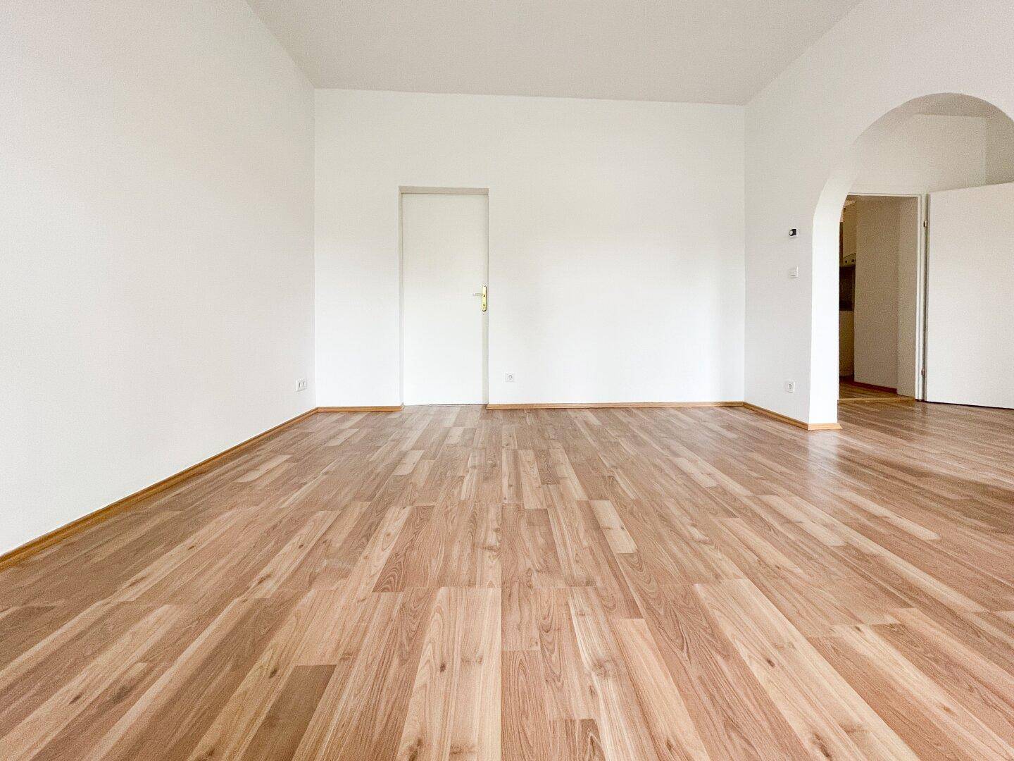 Leeres Zimmer mit Holzboden und weißer Tür, ein Rundbogen führt zu einem angrenzenden Bereich.