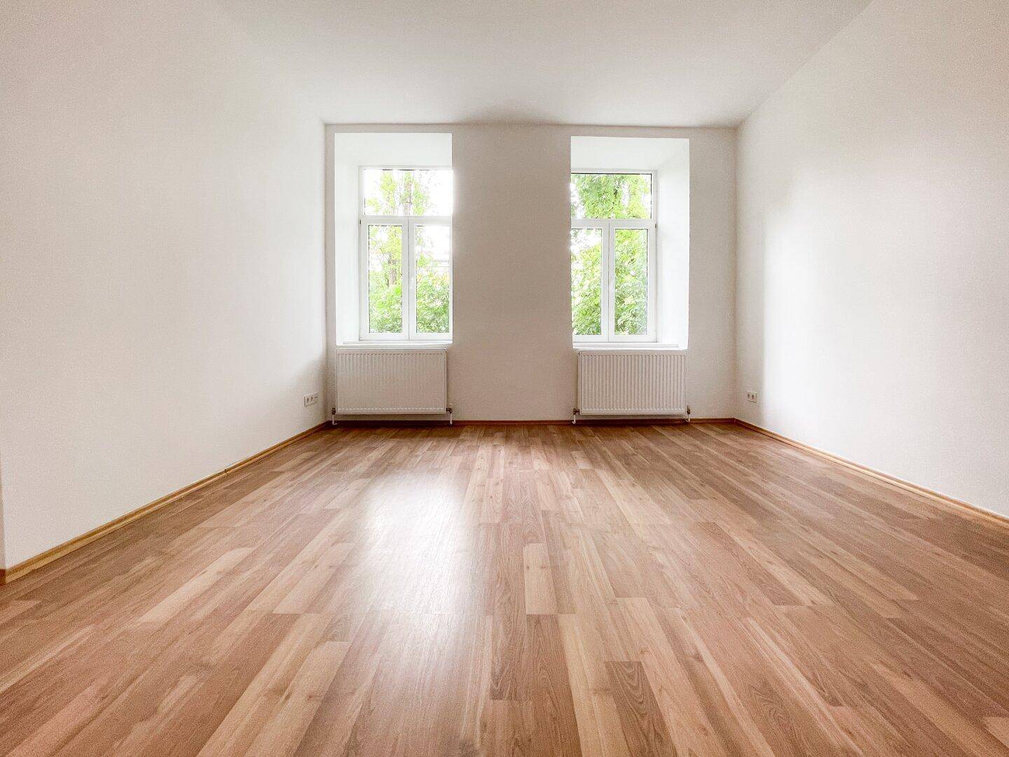 Geräumiges, leeres Zimmer mit Holzboden und zwei großen Fenstern, die viel Tageslicht hereinlassen.