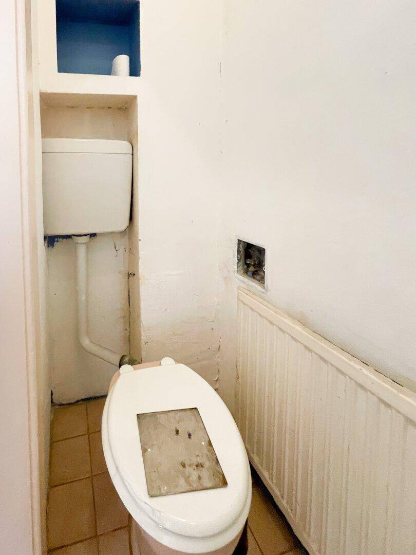 Toilette in einem kleinen Raum mit beigen Fliesen und einem Heizkörper, der eine Modernisierung erfordert.