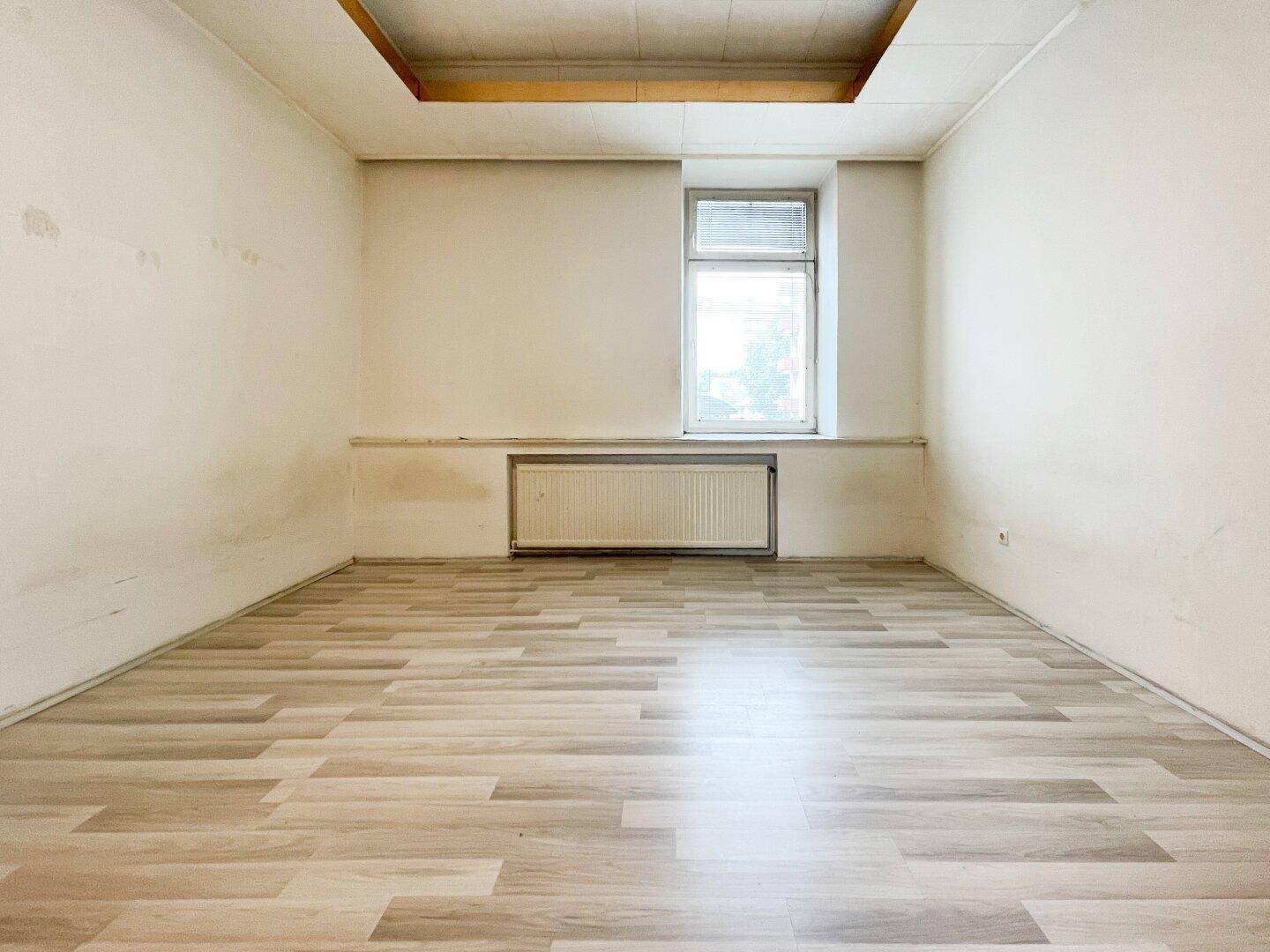 Leerer Raum mit Laminatboden, einem Fenster und einem Heizkörper, bereit zur individuellen Gestaltung.