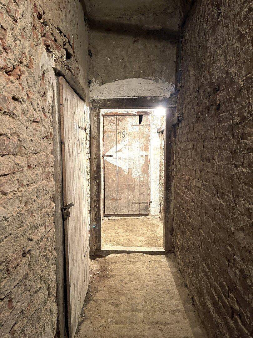 Dunkler, schmaler Kellergang mit unverputzten Ziegelwänden und alten Holztüren.