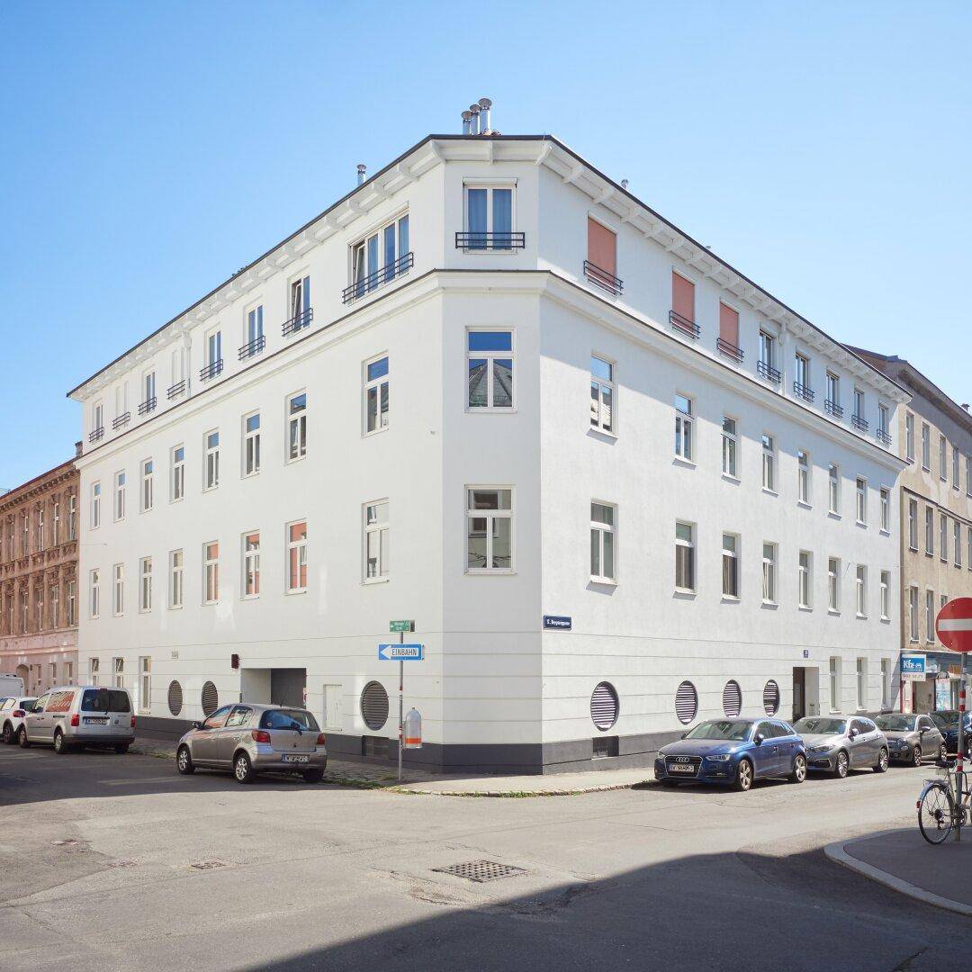 Modernes Eckgebäude mit weißer Fassade, vielen Fenstern und parkenden Autos auf der Straße.