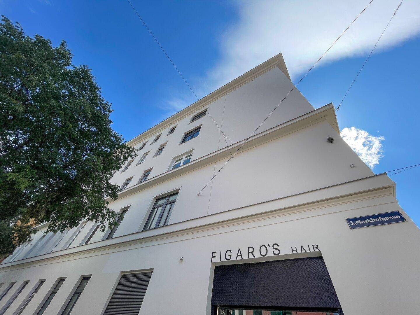 Weiße Fassade eines modernen Gebäudes unter blauem Himmel mit Baum und Straßenschild.