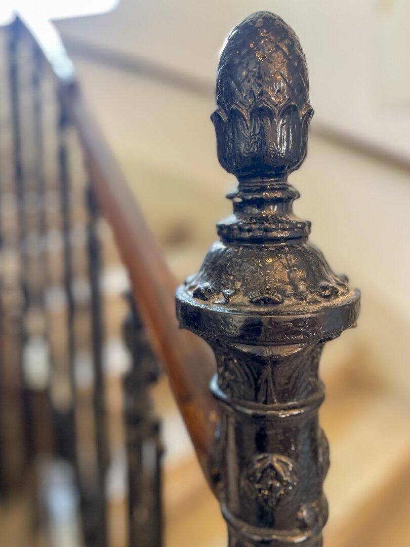Detail eines verzierten Treppengeländers mit dunklem Metallpfosten und Holzhandlauf, das den traditionellen Stil des Gebäudes unterstreicht.