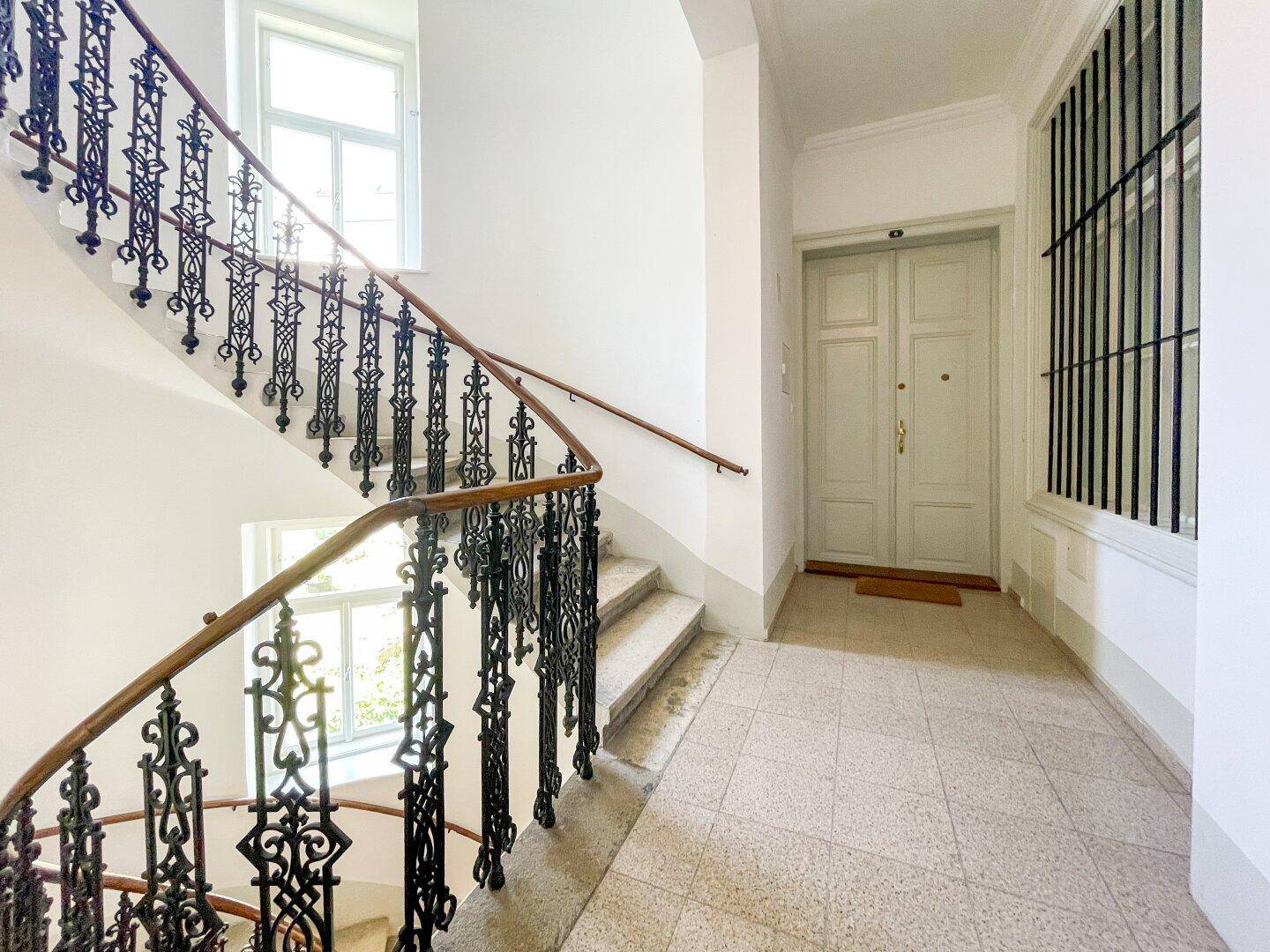 Elegantes Treppenhaus eines Altbaus mit kunstvollem Geländer und großen Fenstern.