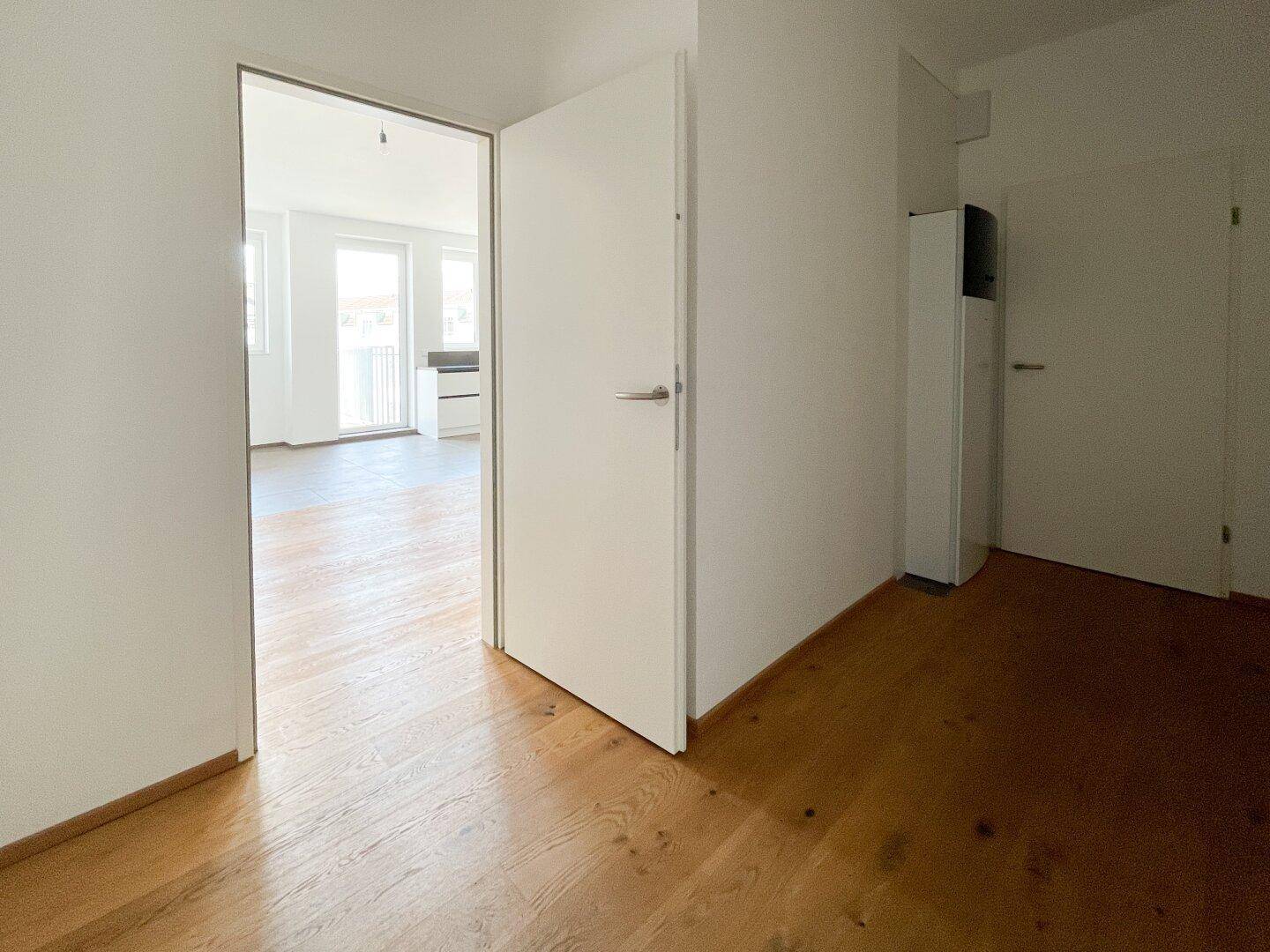 Heller Flur mit Holzboden und weißer Tür, die zum offenen Wohnbereich führt.