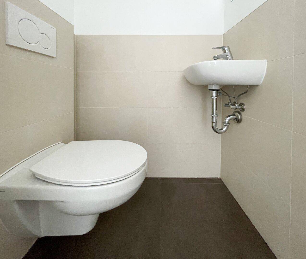 Separate Gästetoilette mit wandhängendem WC und kleinem Waschbecken, in neutralen Farben gefliest.