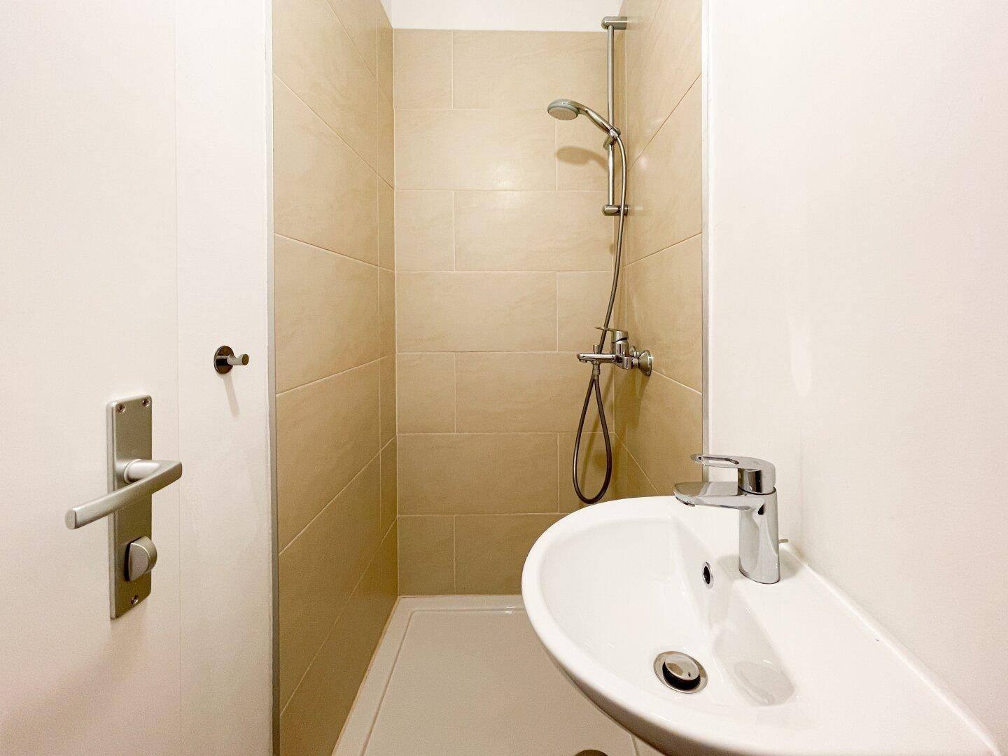 Kleines Badezimmer mit Duschbereich, beigefliesten Wänden und einem einzelnen Waschbecken.