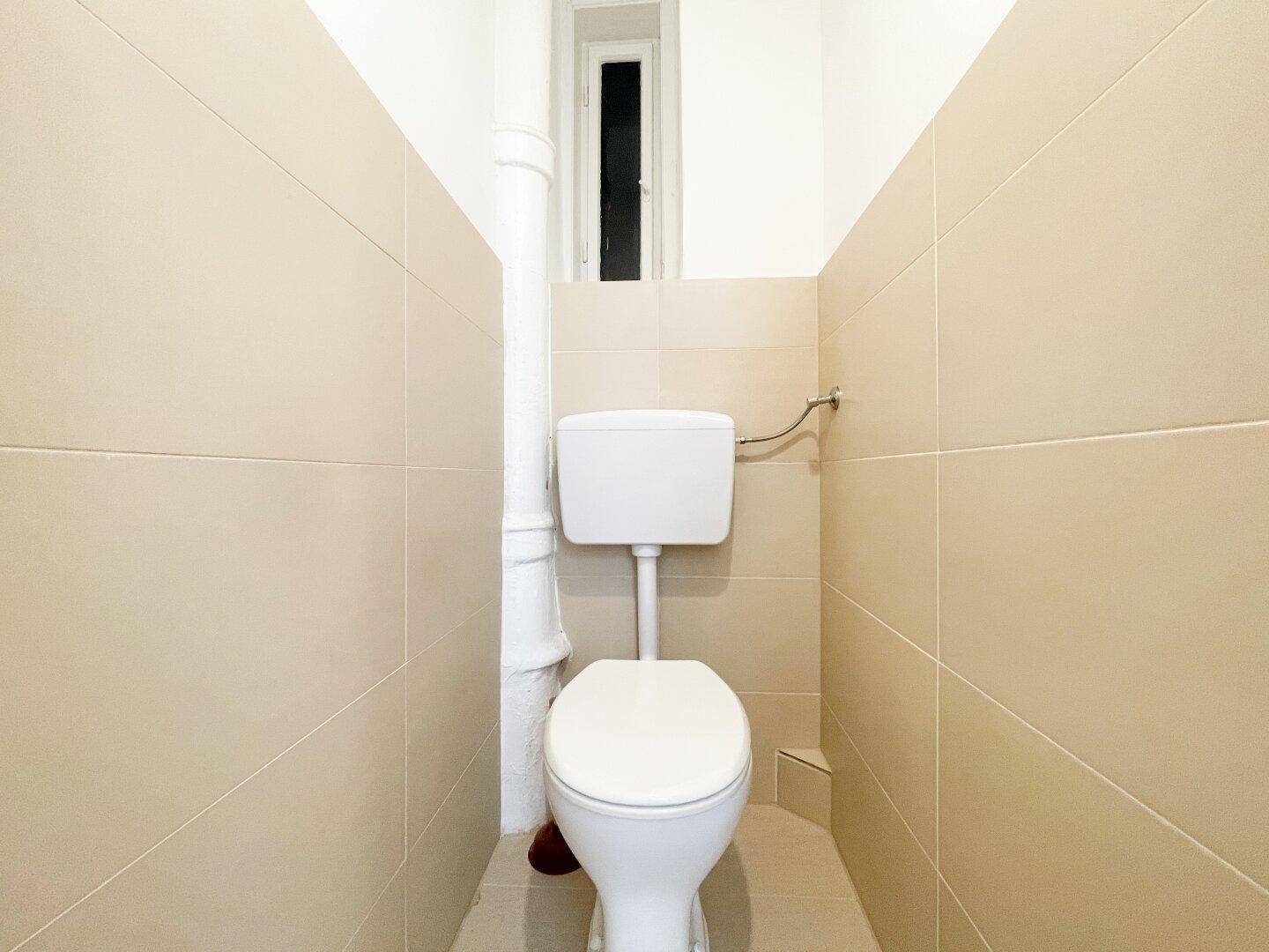 Separates WC mit beigefliesten Wänden, einem Fenster und einer Standardtoilette.