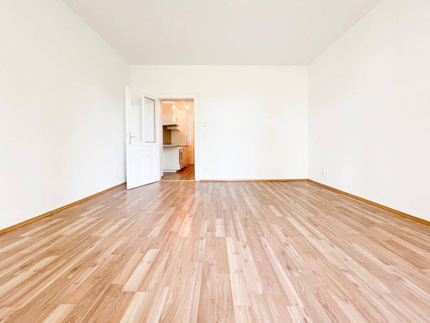 Geräumiges, helles Zimmer mit Holzfußboden und Zugang zu einer angrenzenden Küche.