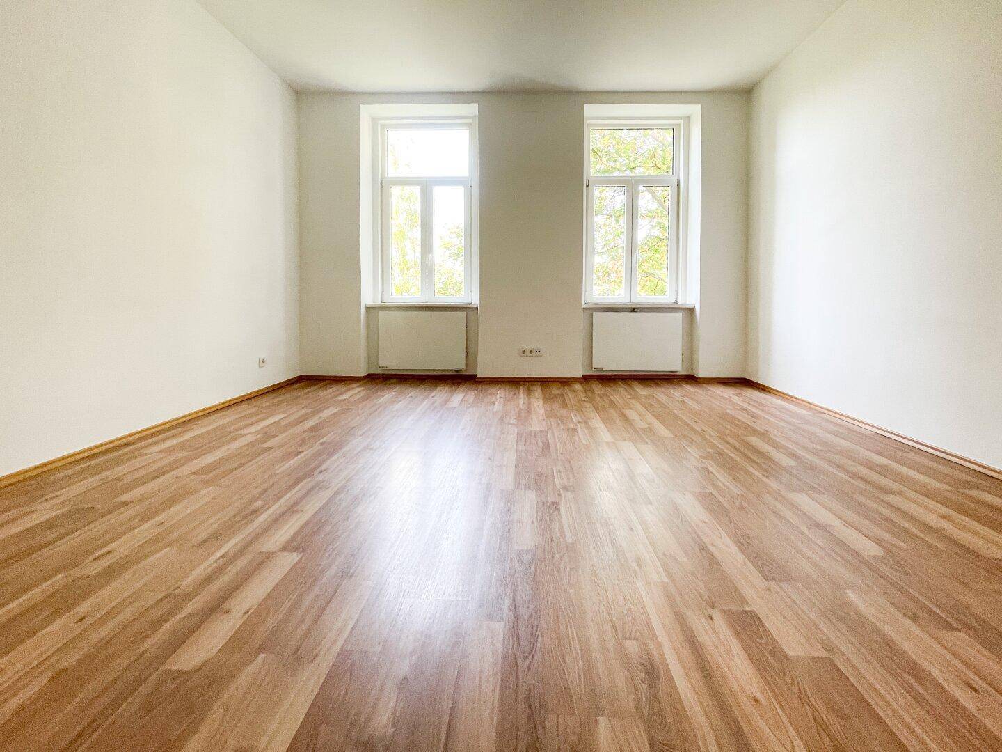 Helles, leeres Zimmer mit zwei großen Fenstern und Holzfußboden, bereit zur individuellen Gestaltung.