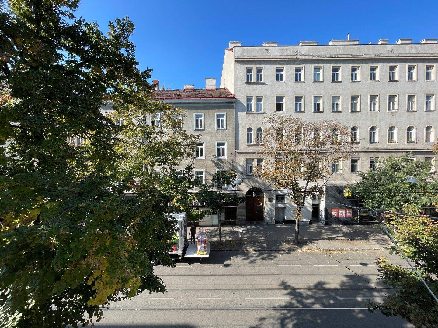 Blick auf die Straße mit Bäumen und gegenüberliegenden Gebäuden an einem sonnigen Tag.