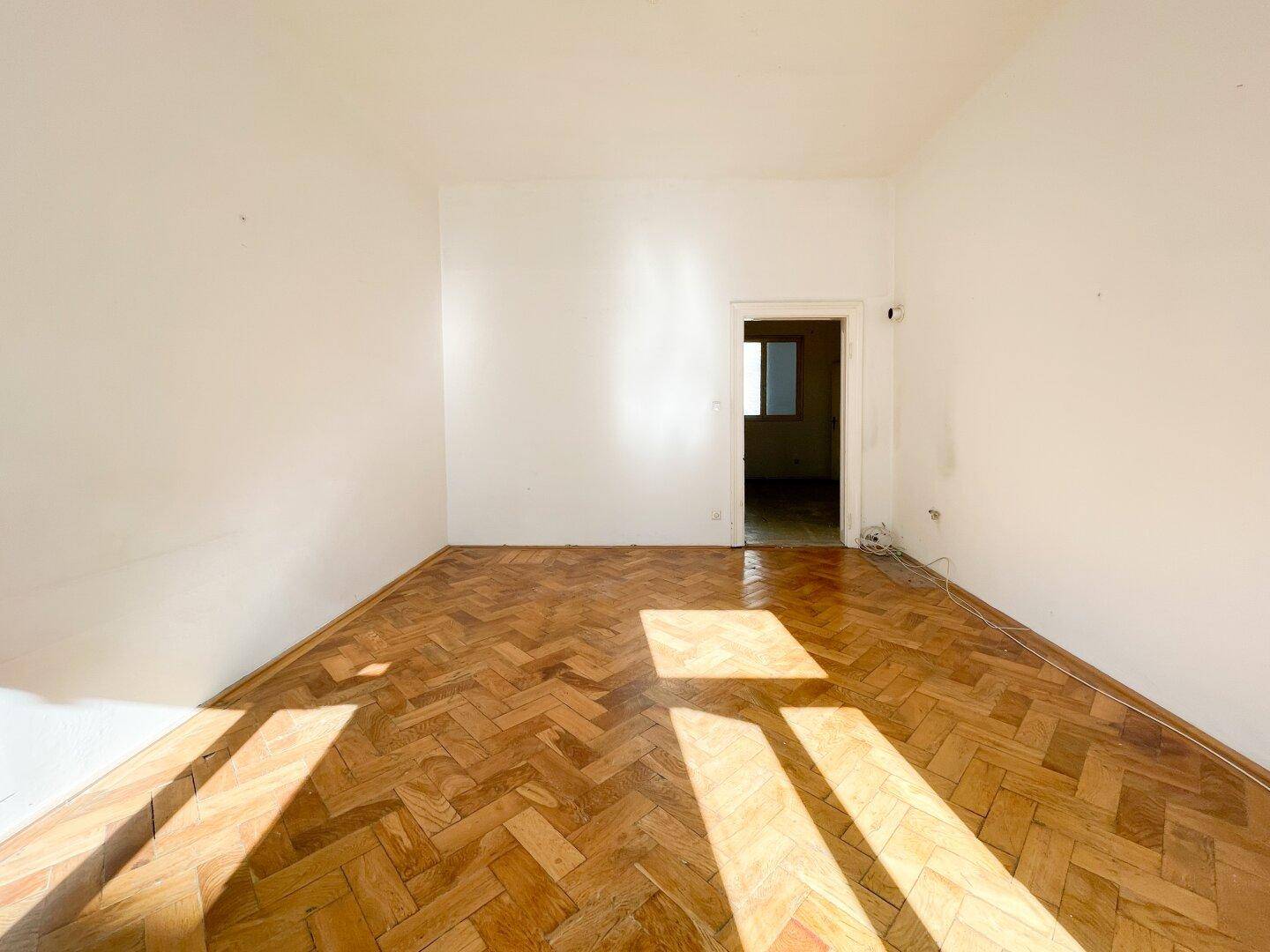 Geräumiges Zimmer mit Fischgrätparkett und Zugang zu einem weiteren Raum, hell und einladend.