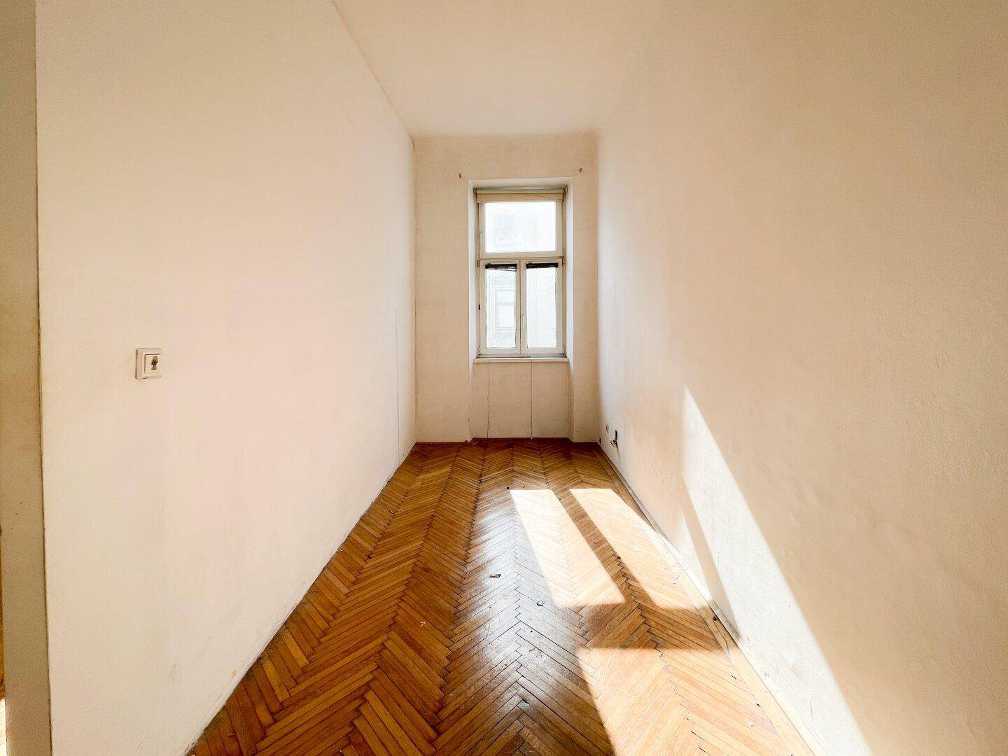 Helles Zimmer mit Fischgrätparkett und einem Fenster, das den Raum mit Sonnenlicht füllt.
