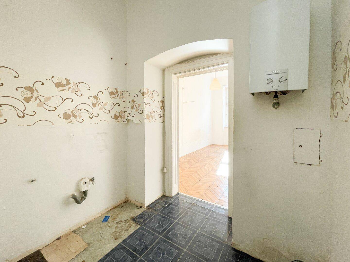 Kleiner Raum mit floraler Tapetenbordüre und Warmwasserbereiter, mit Zugang zu einem weiteren Zimmer.