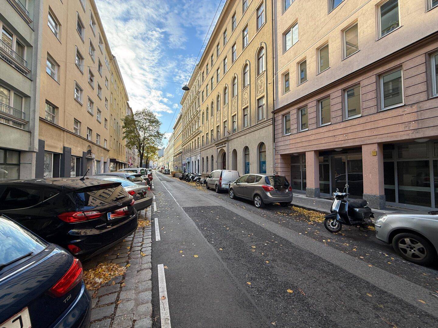 Eine belebte Stadtstraße mit parkenden Autos und hohen Gebäuden unter blauem Himmel.