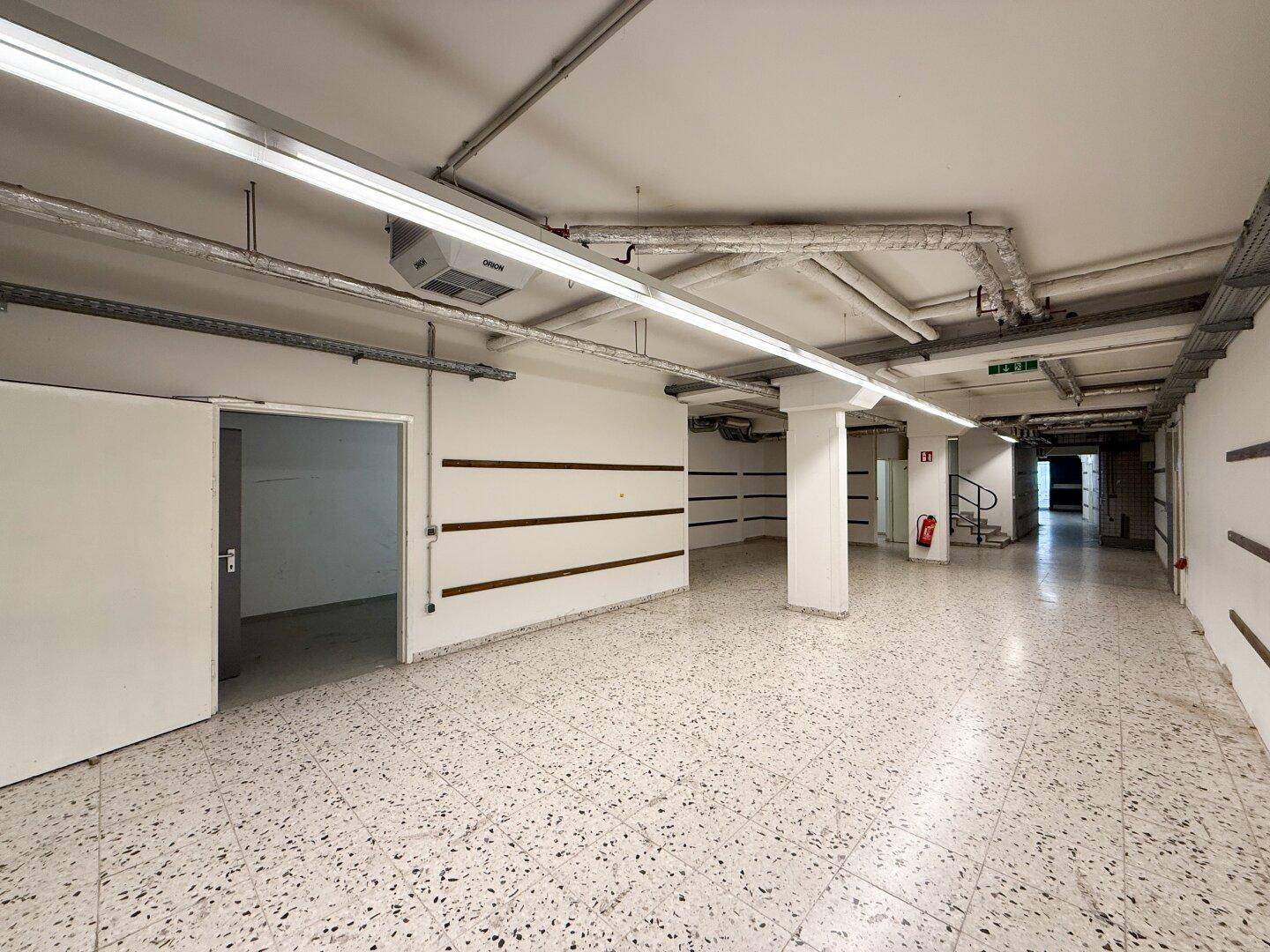 Großer Kellerraum mit Terrazzoboden, freiliegenden Rohren und Zugang zu weiteren Bereichen.