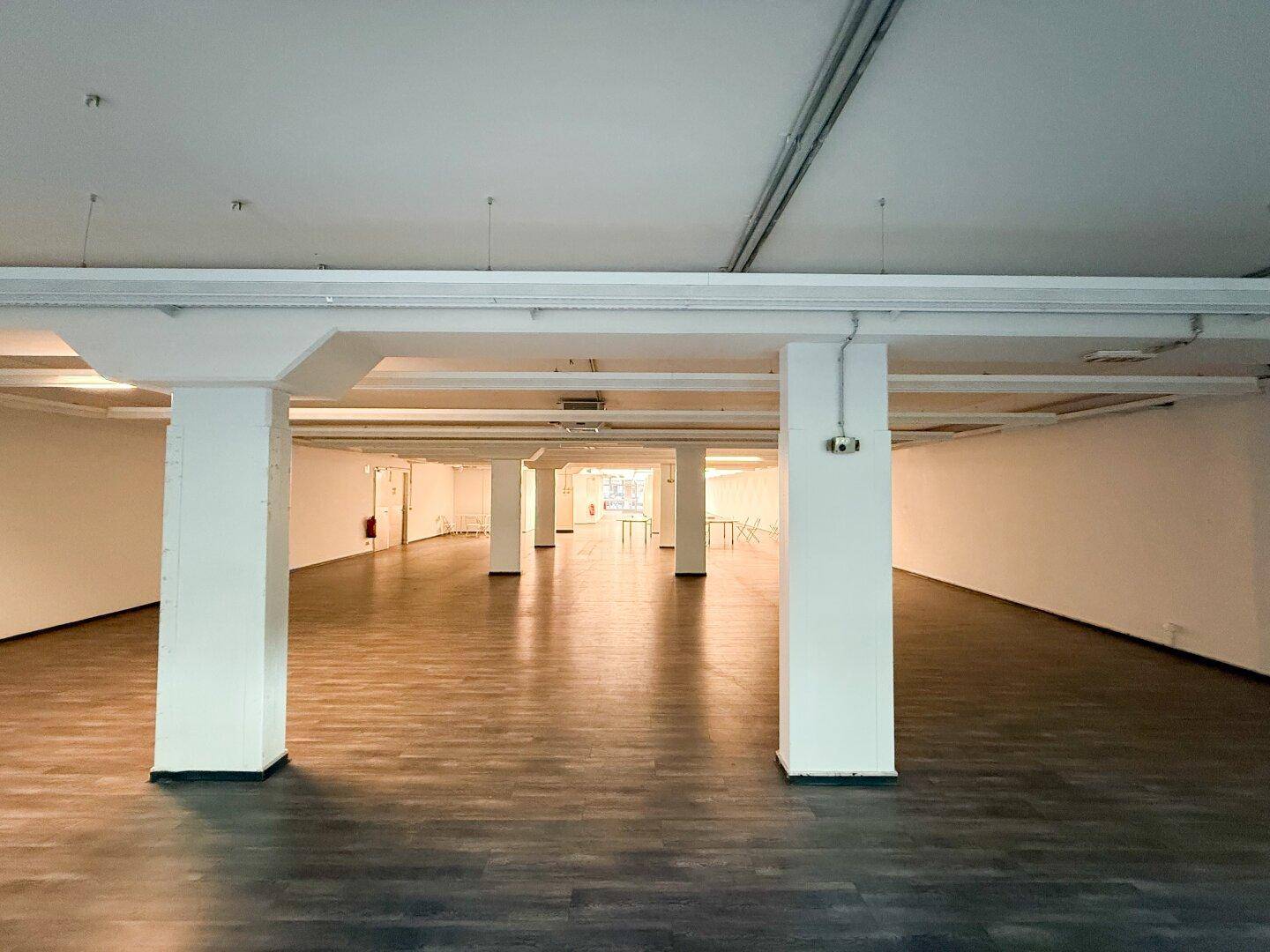 Langer, offener Gewerberaum mit Säulen und dunklem Holzoptik-Laminatboden.