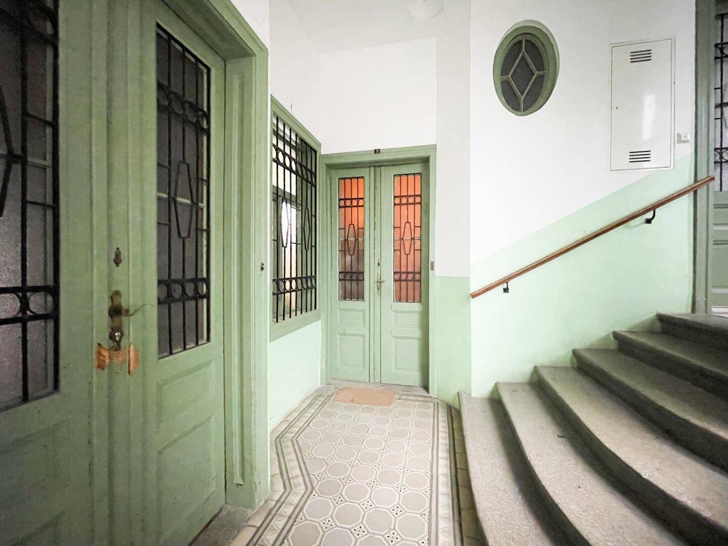 Historischer Eingangsbereich mit gemusterten Fliesen, grünen Wänden und einer Treppe mit Holzgeländer.